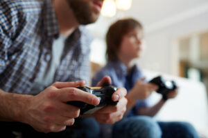 Young man with joystick playing video game on background of his son