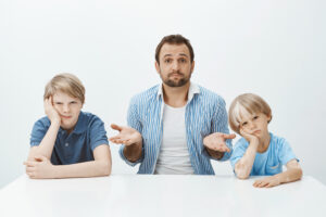 Portrait of questioned good-looking european father sitting at table with bored and upset sons, shrugging with raised palms, being clueless how to raise boys alone, sitting confused over grey wall.