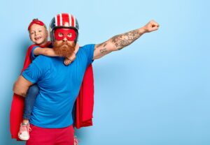 Powerful dad gives piggy back to child, demonstrates courage, makes flying gesture, wears helmet, red mask, cape, have fun together, stand against blue wall with empty space, brave help someone else