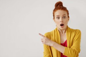 Surprise and astonishment concept. Human feelings and reaction. Portrait of shocked amazed young Caucasian ginger woman pointing index finger sideways, indicating blank copyspace wall for your text