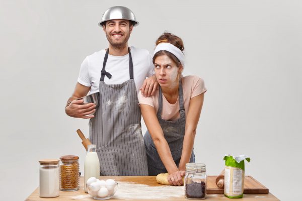 Exhausted woman with flour on face, kneads dough, tired of preparing homemade bread, cheerful man in apron leans on shoulder, holds bowl, glad to help wife on kitchen. Couple make pizza together