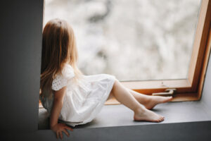 Little child in tender white dress is sitting on the windowsill, kid, child services, happy childhood, lonely tiny girl